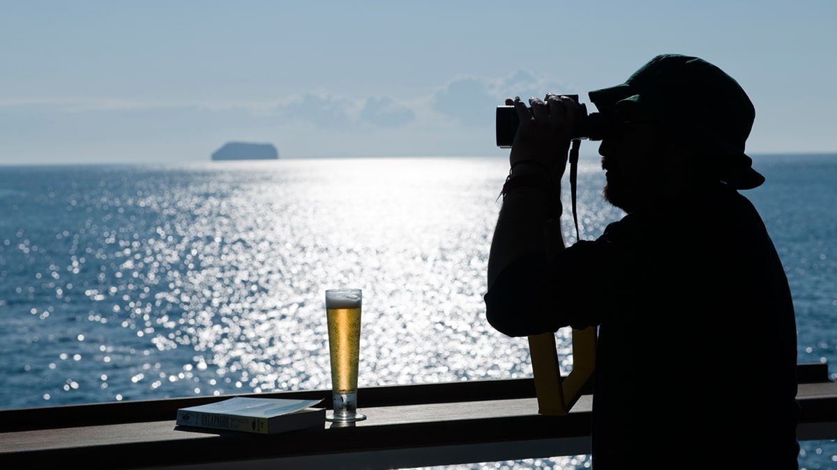 Un voyageur observe l'horizon depuis un luxueux bateau de croisière, savourant une bière, avec des paysages côtiers à couper le souffle. TMR Croisières.